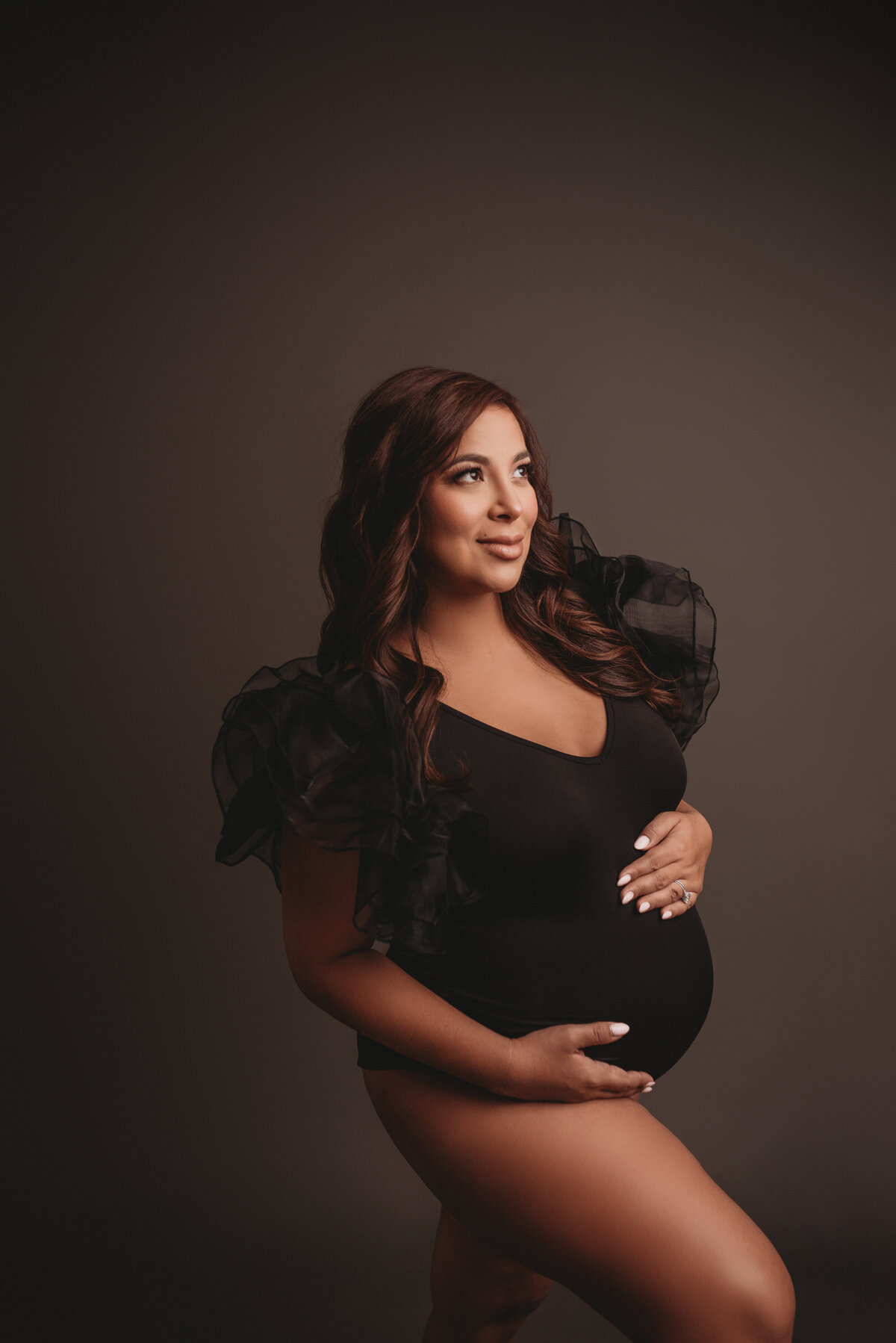 Pregnant woman at maternity and newborn studio of Atlanta posing for maternity portrait wearing black ruffle body suit standing up holding baby bump