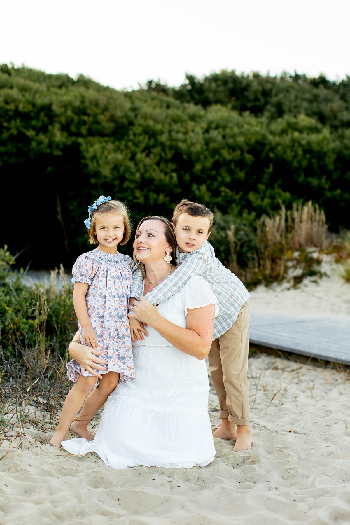 A mother in a white dress embraces her two smiling children at golden hour on the beach in Virginia, captured in a candid and heartfelt moment by Brooke Tucker photography