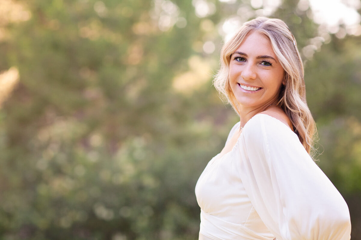 A woman with long blonde hair smiles while standing outdoors. She is wearing a light-colored top, and the background features blurred greenery, suggesting a natural, sunny setting.