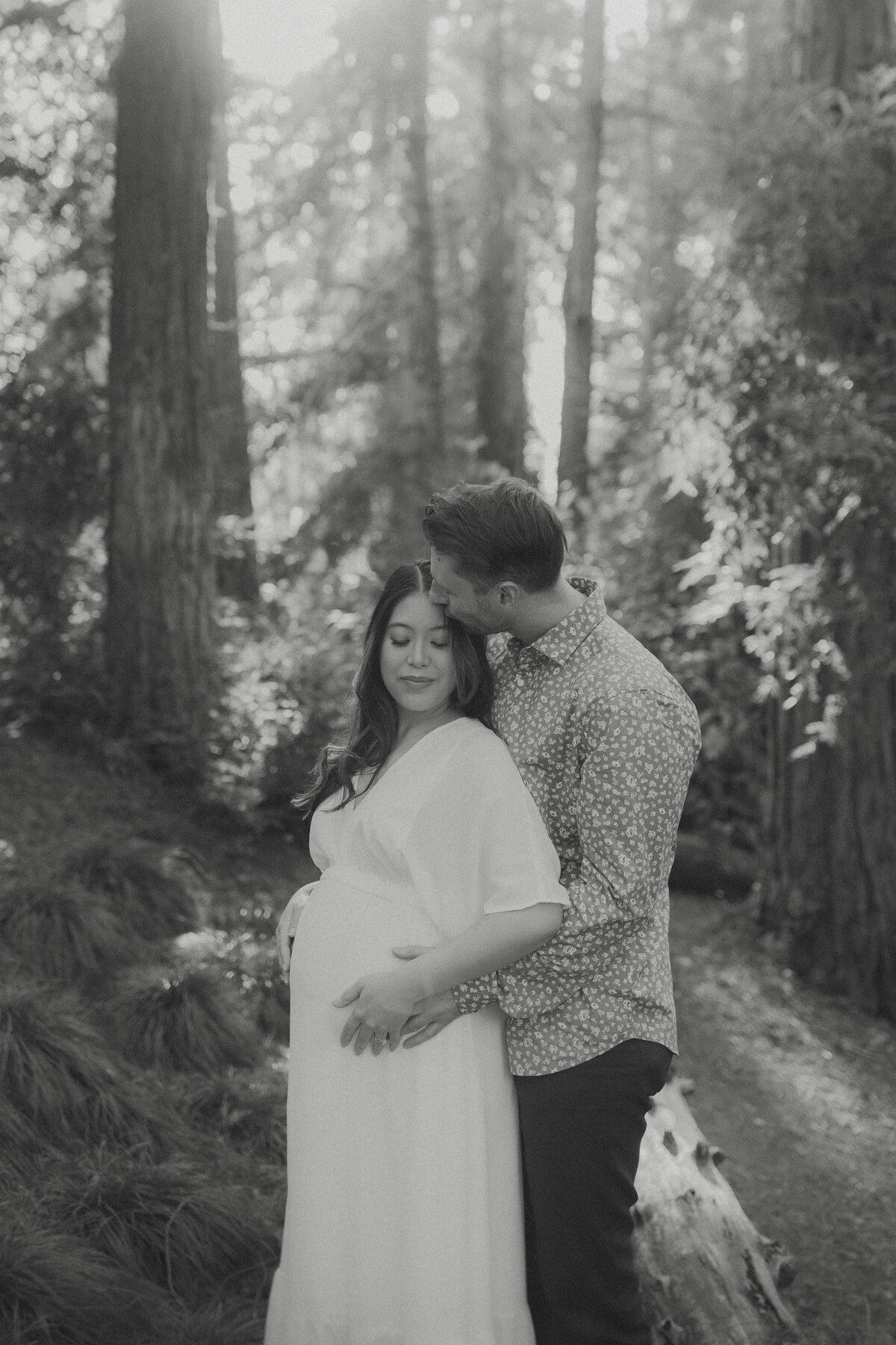 A mother-to-be embraced by her partner in a serene forest setting, captured in an elegant and documentary maternity photography style.