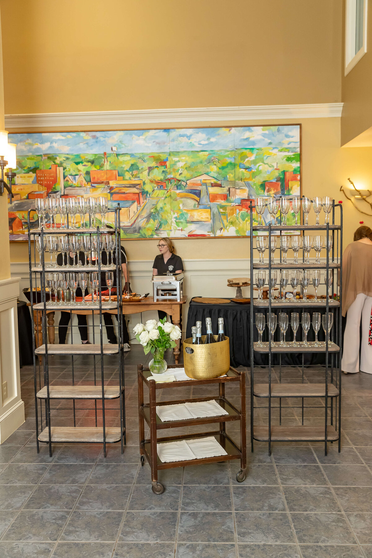 A drink station with racks of empty wine glasses, a cart with a gold ice bucket and bottles, and a person standing behind a table. A colorful painting hangs on the wall in the background.