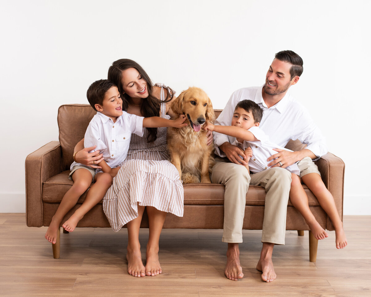 Family dressed in all white sitting on a couch petting their dog pose candidly for their family photo