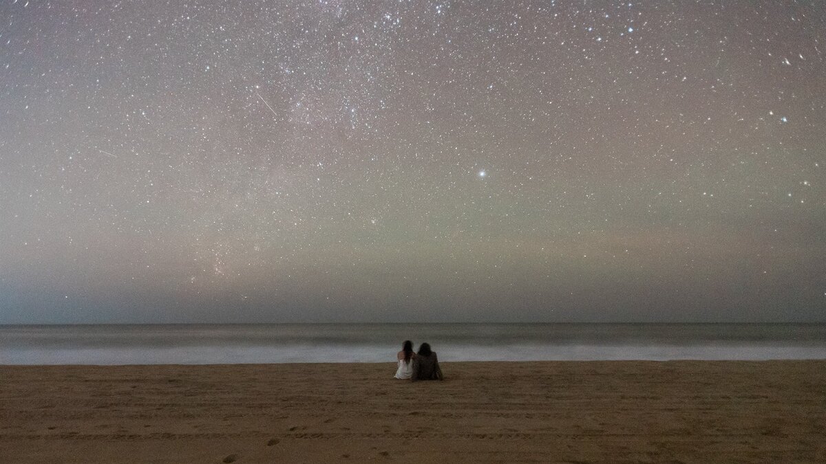 A bride and groom in their wedding finest sit on the sandy beach in the dark night during their adventurous luxury Oaxaca Mexico Elopement.  The night sky is littered with stars and in front of them, a milky white bioluminescent wave crashes ashore. 