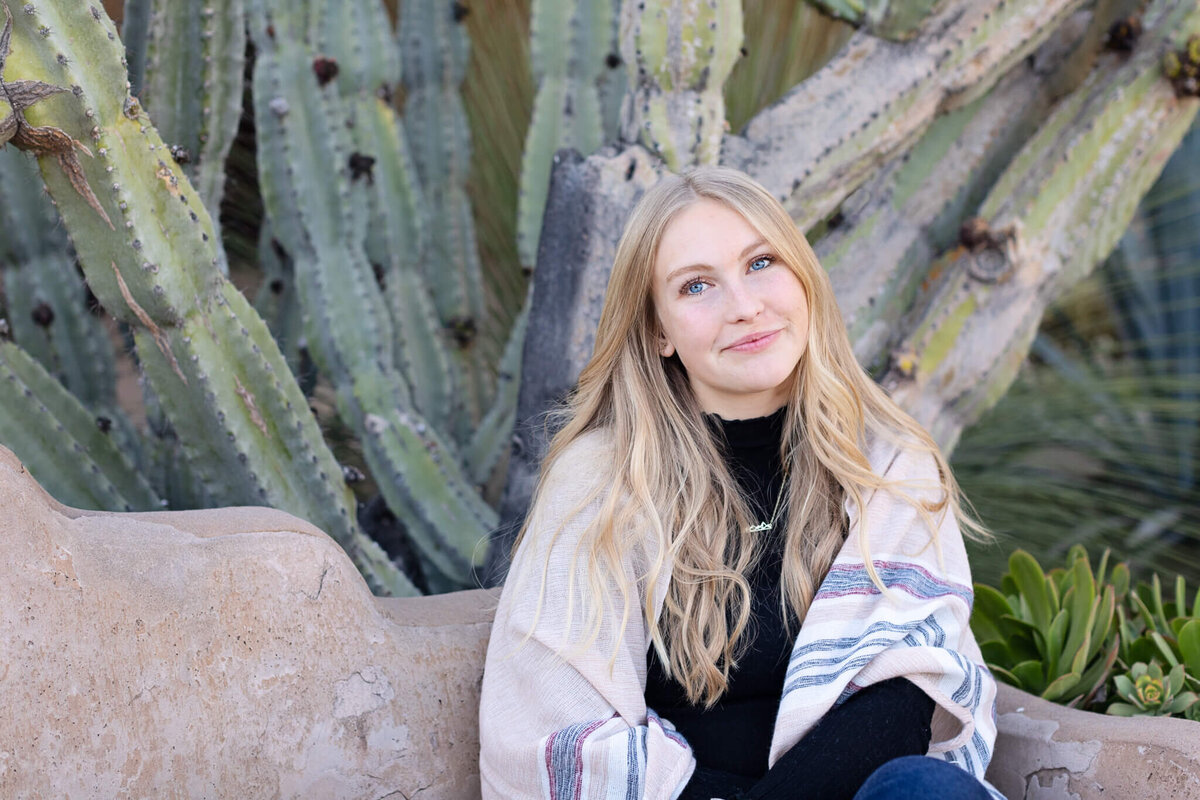 A young woman with long, blonde hair sitting on a bench wearing a black shirt and white and gray shawl over her shoulders.