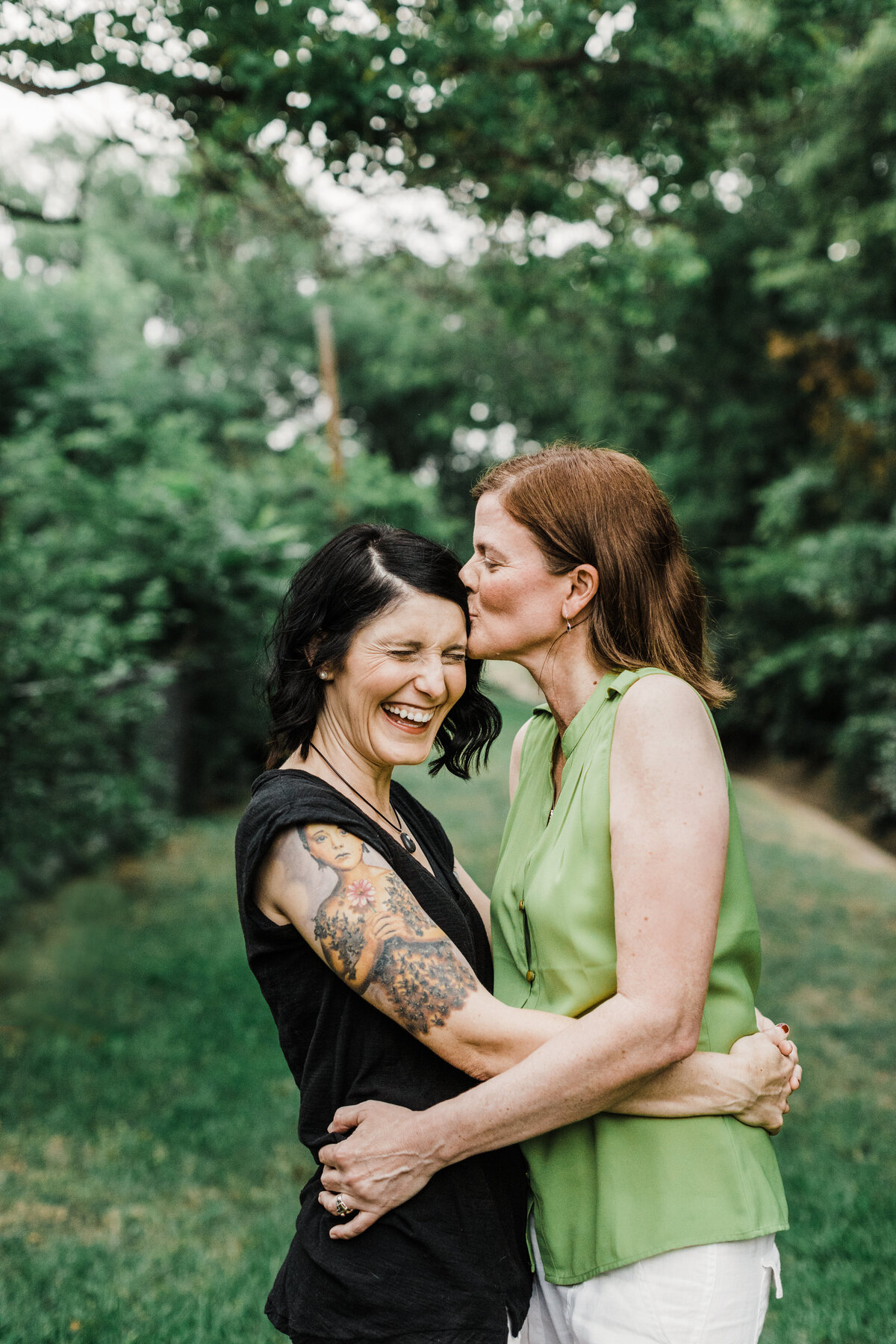 Two women holding each other close with one laughing while the other gives her a kiss on the forehead during their engagement session in DFW, Texas. The woman on the right is wearing a sleeveless green blouse and white pants while the woman on the left is wearing a sleeveless black blouse and has a half sleeve of tattoos on her arm. They are backed by many trees and other greenery.