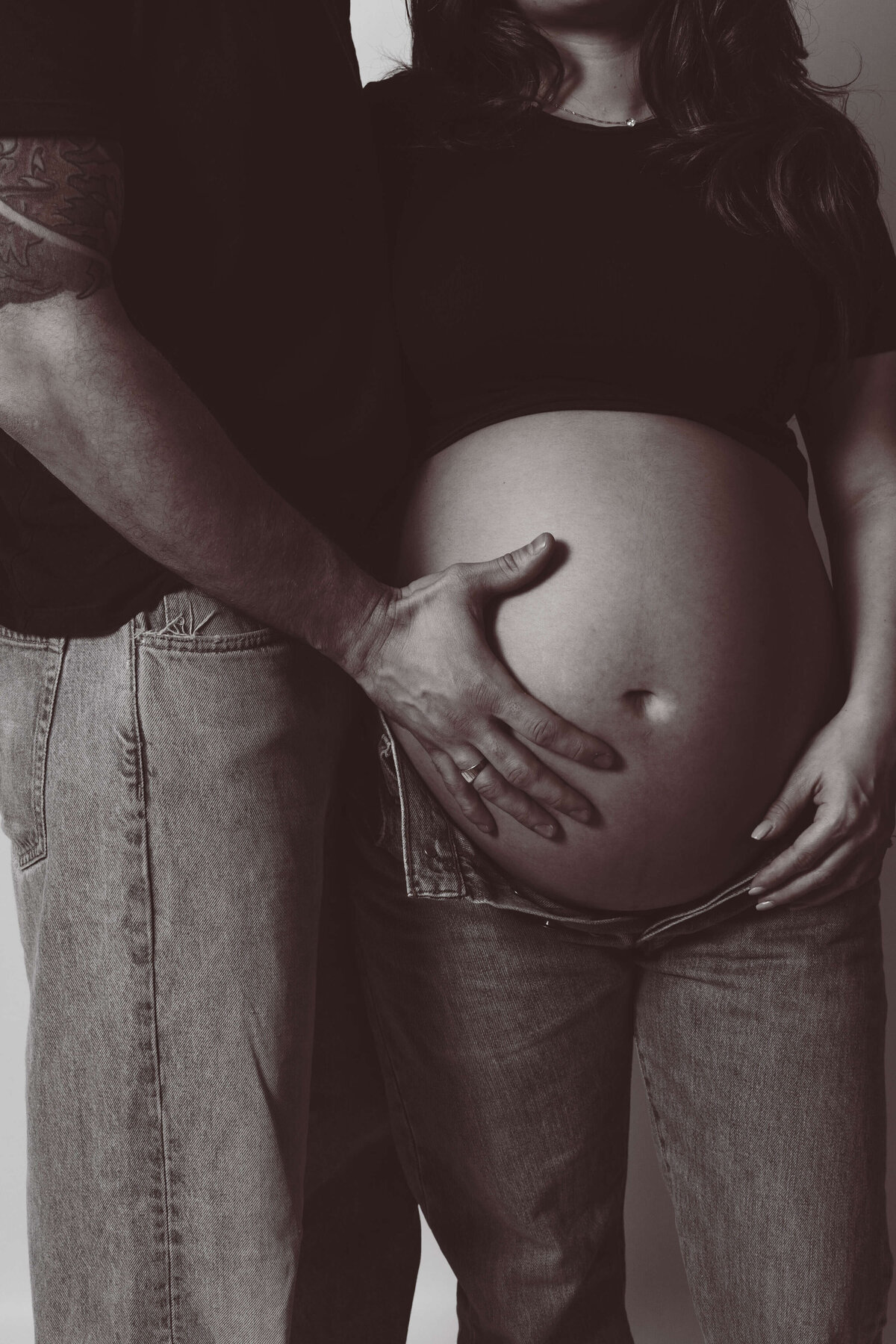 Närbild på en gravid kvinna ståendes bredvid hennes partner som har en hand på hennes gravida mage under en gravidfotografering i studio i Stockholm