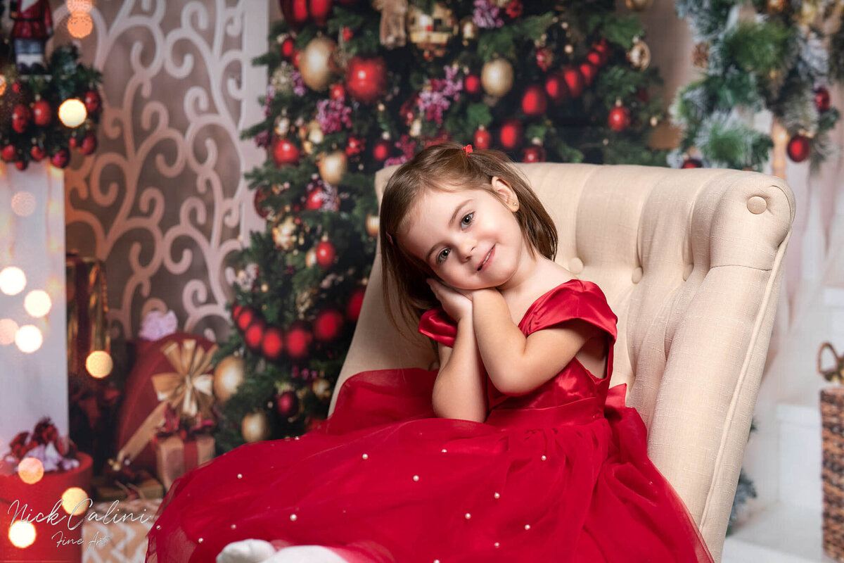 Little girl in red dress posing on beige armchair in Calini Weddings Studio - elegant Christmas tree and festive bokeh lights.