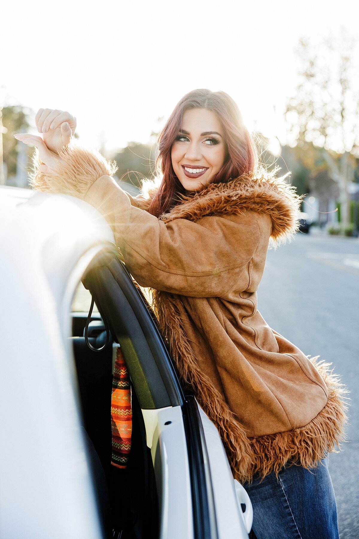 Sun-kissed senior portrait of a teen girl smiling by a car in a fur-trimmed suede jacket, shot in golden hour light in Santa Clarita