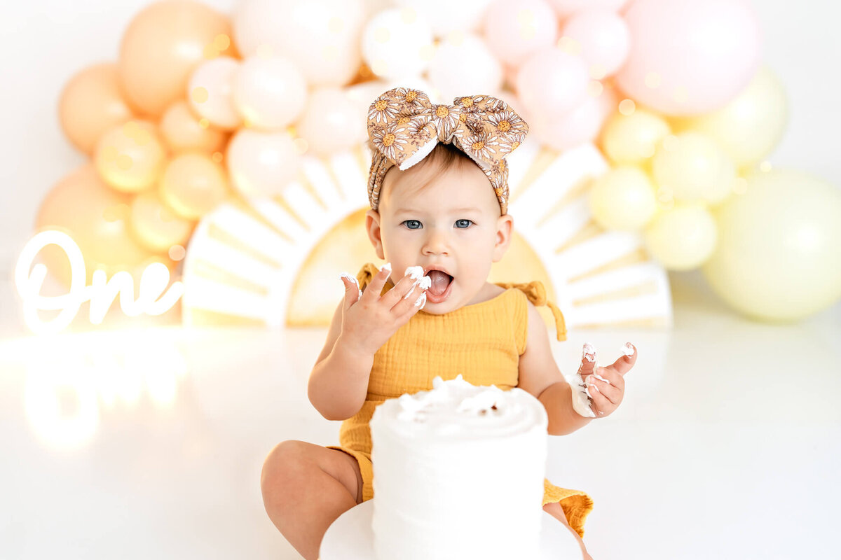 A toddler in a yellow outfit sits in front of a white cake, holding some frosting. Behind her, there are pink and peach balloons and a "One" sign, creating a festive atmosphere. She has a big, patterned bow on her head.