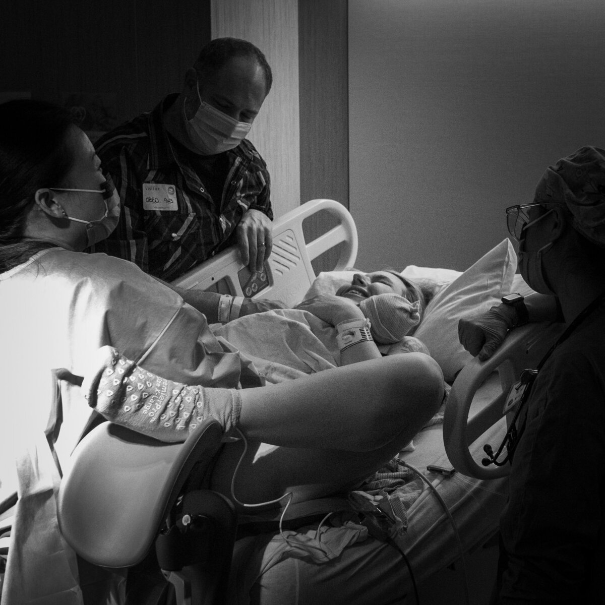 a woman is laying down in a hospital bed after giving birth. Her legs are spread and the doctor is standing between them helping to deliver the placenta. The new dad and nurse stand at her sides surrounding the birthing woman.
