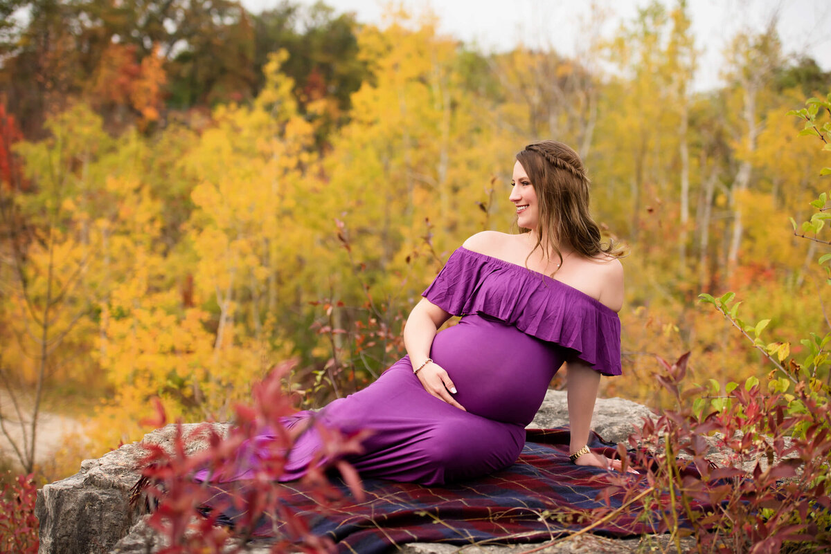 Pregnant woman wearing a purple dress sitting on rock with fall foliage behind her for her Burlington, Ontario outdoor maternity photography session