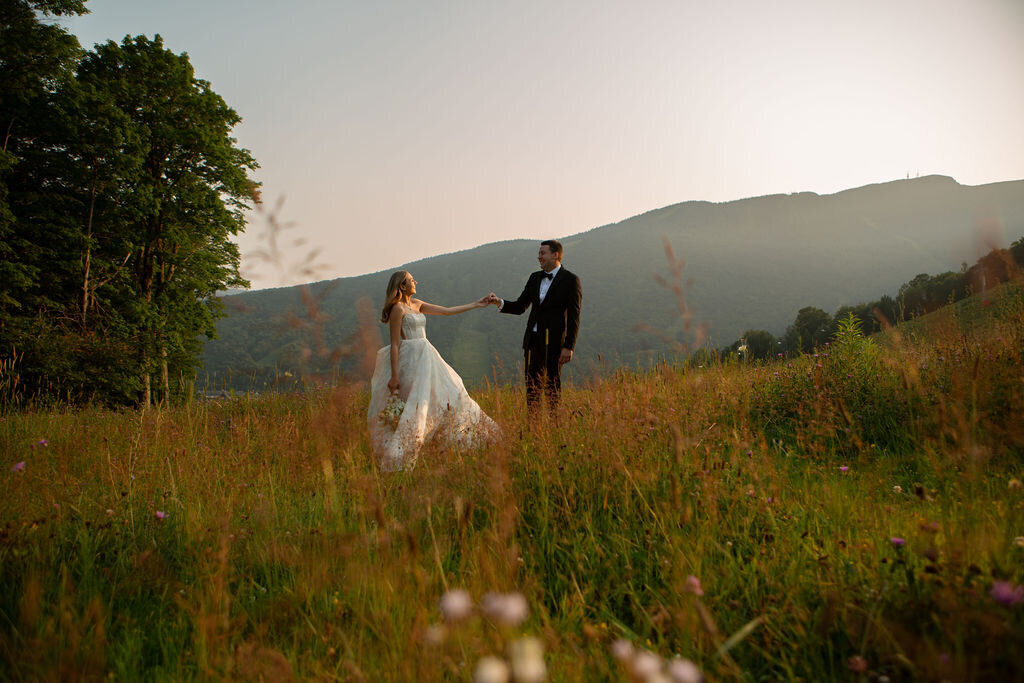 A bride and groom outside at their wedding in Vermont. Standing in a field and holding hands with the mountains in the background.