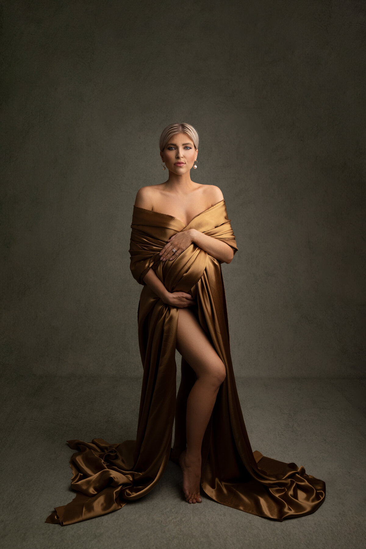 Woman poses for fine art maternity photos with Philadelphia Main Line's best maternity photographer Katie Marshall. Woman in a gold silk photoshoot gown is standing facing the camera with the train of the dress puddled at her feet. Her leg is bent and peeking out from the space where the gown wraps. One hand is resting underneath her bump, the other overtop. She is looking intently at the camera. Full body image.