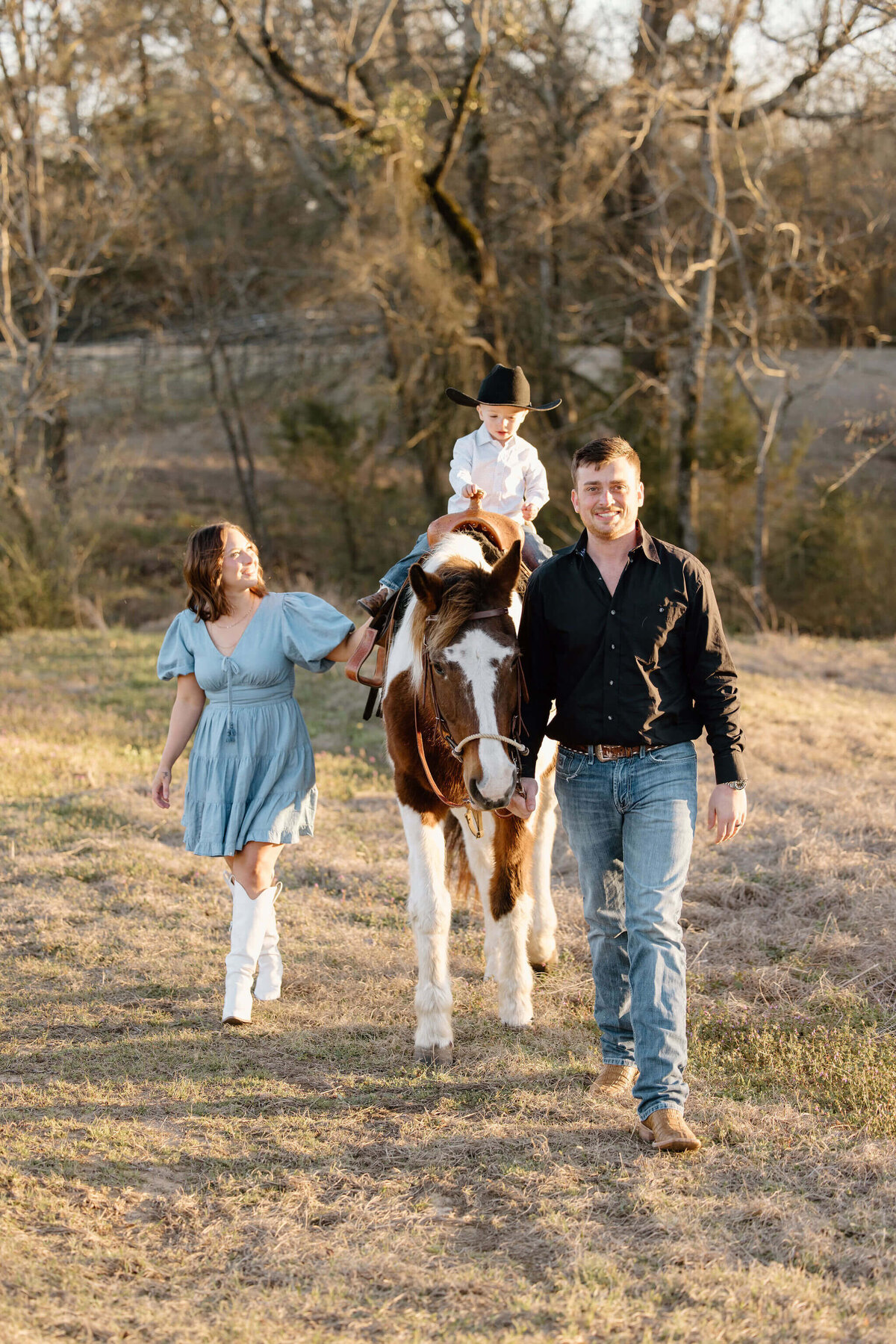 young family in denim and white walking through East Texas pasture with son on horse