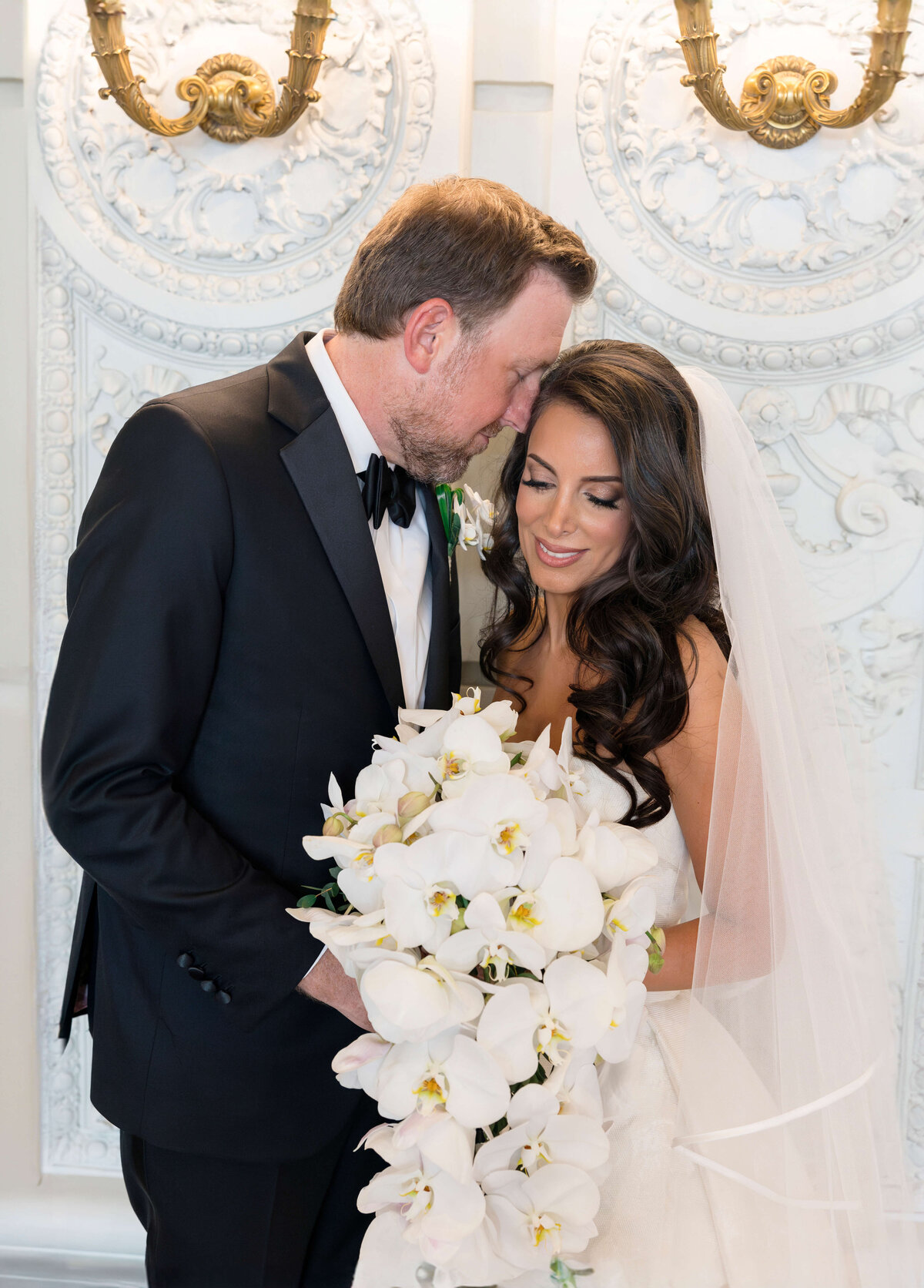 A groom in a black tuxedo tenderly leans to kiss his bride on the forehead. The bride, wearing a white gown and veil, holds a cascading bouquet of white orchids. They stand against an ornate, white decorative background.