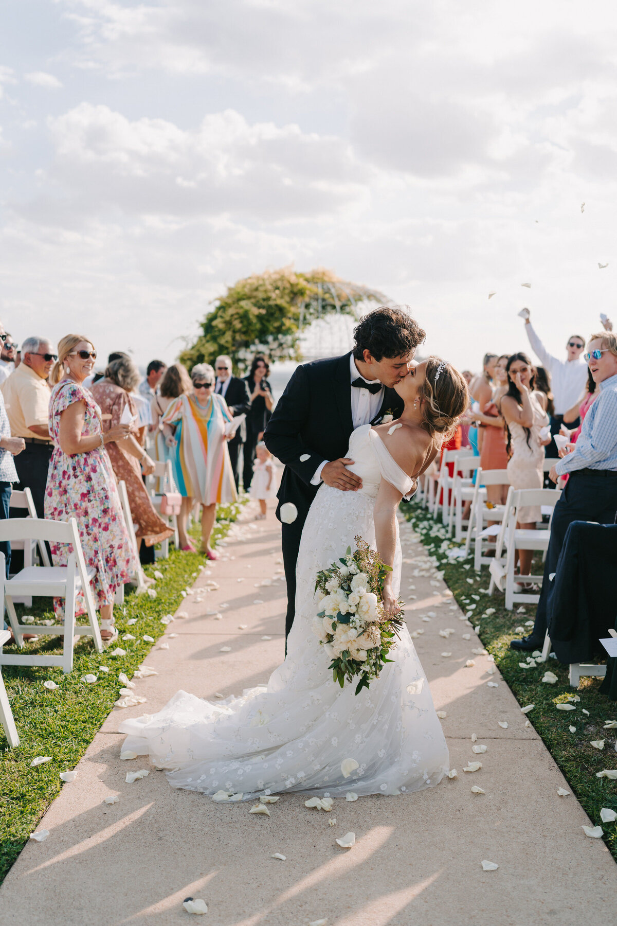 A colorful photograph of Calista and Augustine on their wedding day at Vintage Villas in Austin, Texas. Bride and groom kiss in the middle of the ceremony aisle as she dips her back and holds her white floral bouquet at her side. Guests are turned to look at them, cheering, smiling and throwing white rose petals over them. Wedding photography by Stacie McChesney of Vitae Weddings.