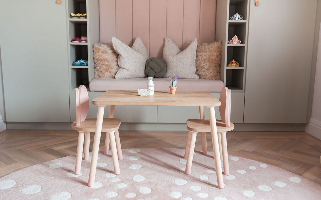 A cozy children's playroom with pastel-colored decor. It features a built-in cushioned seating nook with shelves and a wooden table with pink chairs.