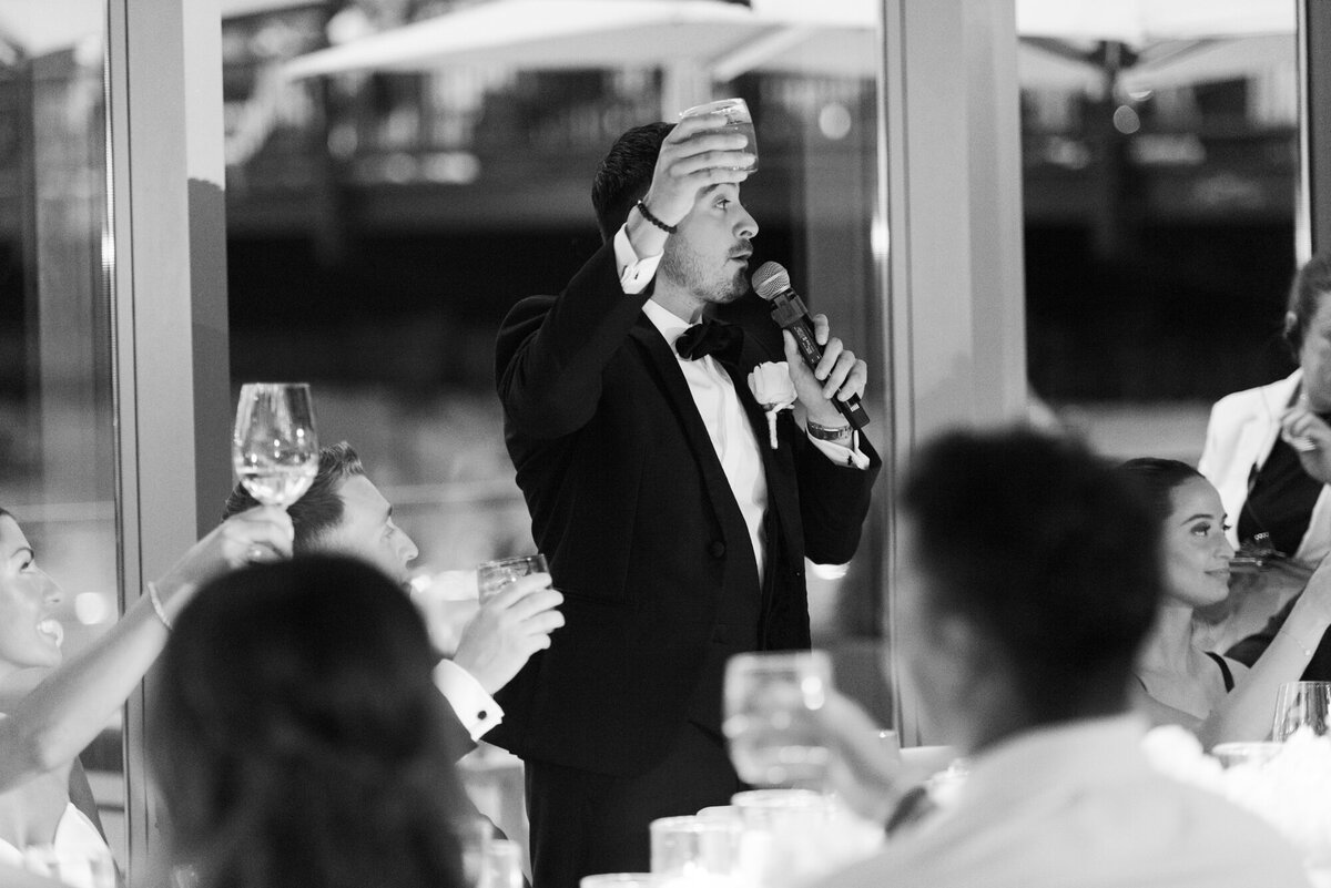 A cinematic black and white photo of a best man raising his glass to give a heartfelt toast at a wedding reception.
