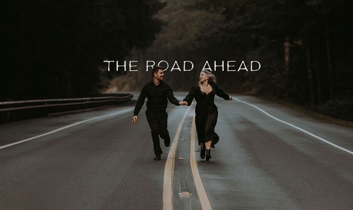 A wedding couple dressed in black running down a road towards the camera, looking at each other and laughing.