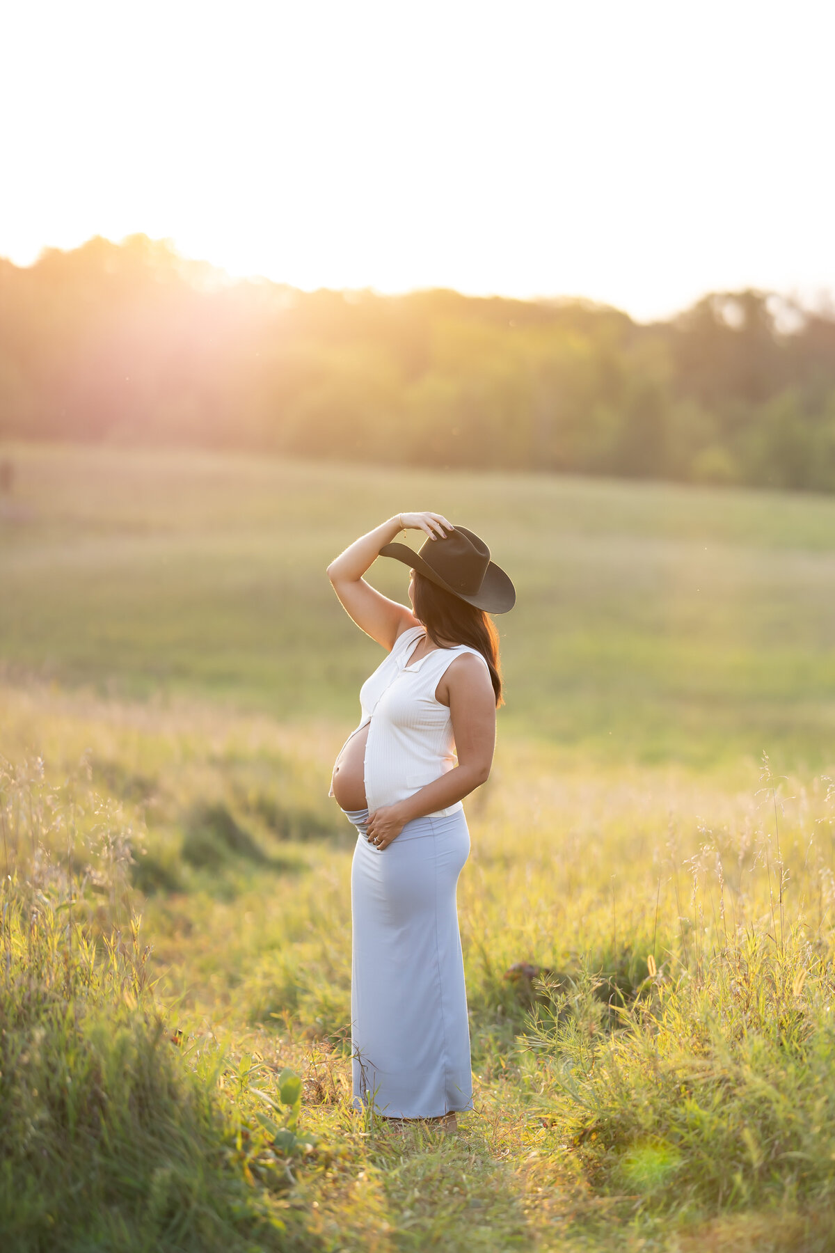 a woman wears a cowboy hat, blue skirt, and white crop top as she holds her baby bump and looks out to the sunset behind her. she stands in a grassy field