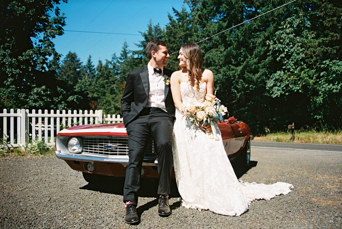 A bride and groom lean against a red convertible car.