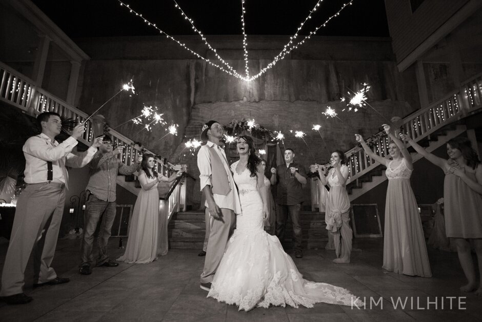 A bride and groom smile and walk hand in hand outside a building at night, surrounded by guests holding lit sparklers and celebrating their wedding.