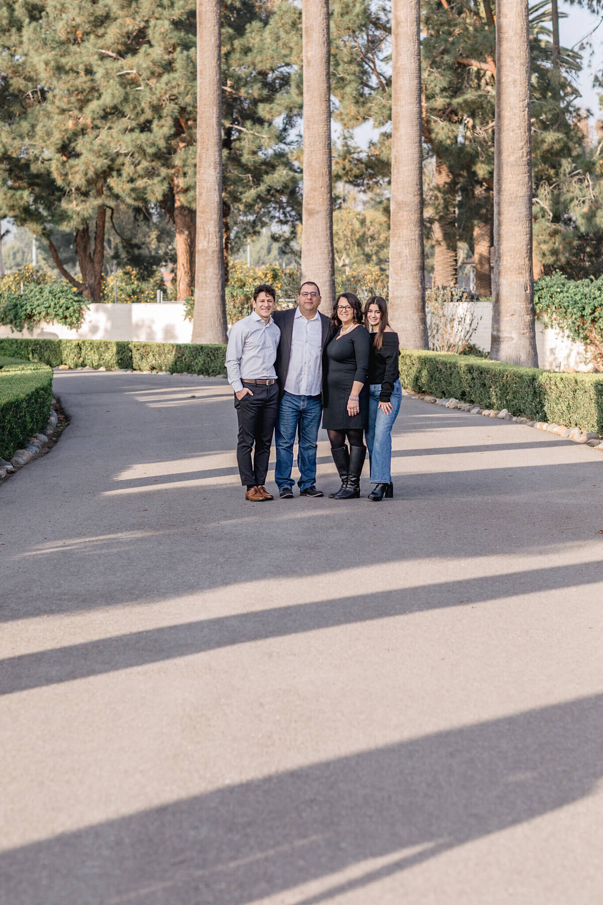 A family of four standing far away at the end of an asphalt path, hugging each other and looking at the camera, surrounded by tall palm trees.
