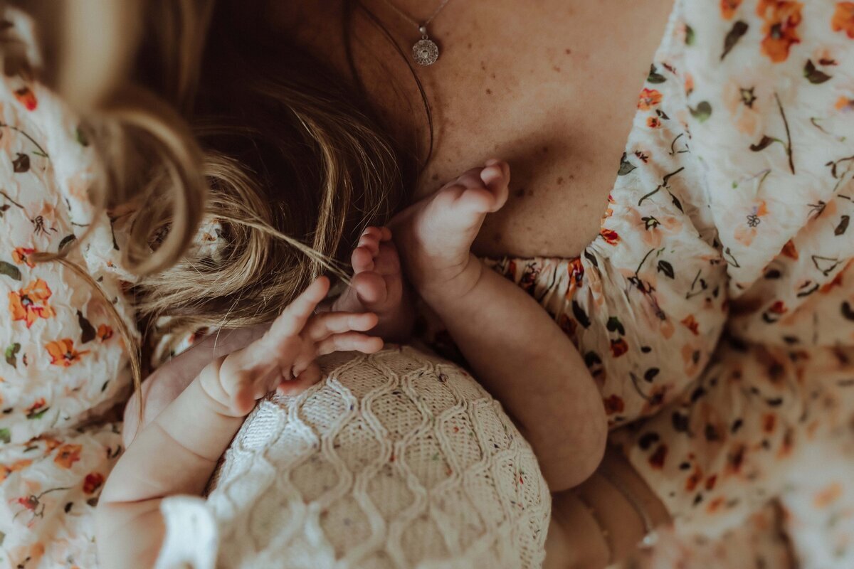 An infant lies on a floral-patterned fabric, with feet extended and hands grasping a knit blanket.