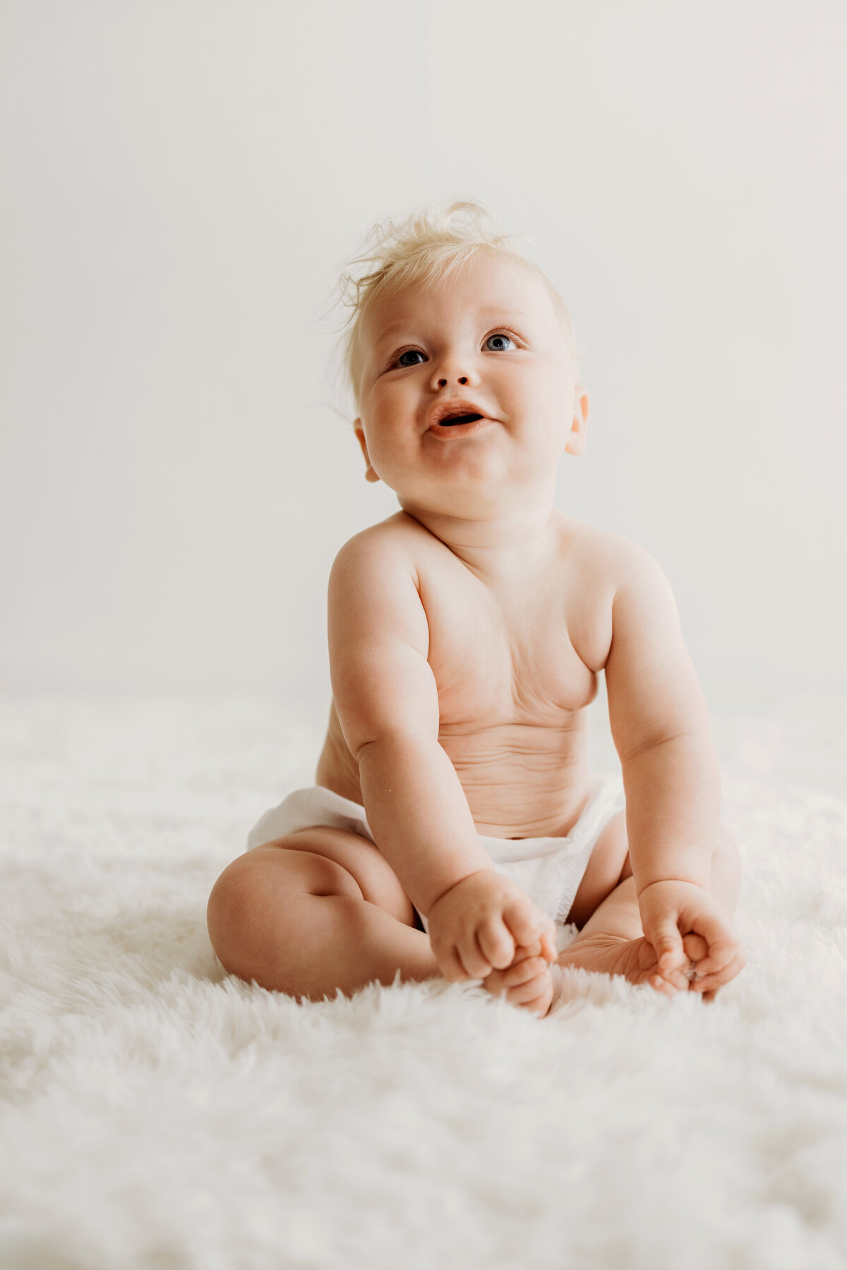 A happy six month old boy sits up nice and tall on a white rug in a white diaper cover for his milestone photos in Denver. 