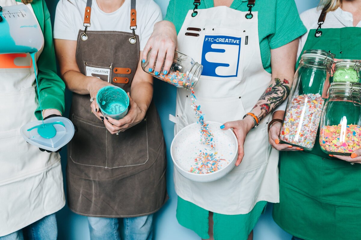 Four people wearing aprons hold jars of sprinkles and paint.
