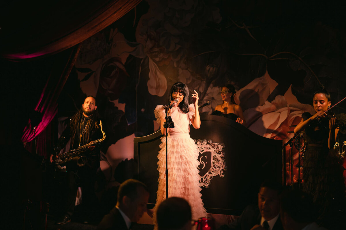 At the tented wedding in Virginia captured by photographer Elisabetta Marzetti, the singer in a white dress took the stage with a vintage microphone. She was gracefully accompanied by musicians playing a saxophone, cello, and a piano. The overall setting was enhanced by a beautiful floral mural in the background, and the soft, dim lighting added to the intimate atmosphere of the performance. Clayton Films also captured the event beautifully as the videographers.