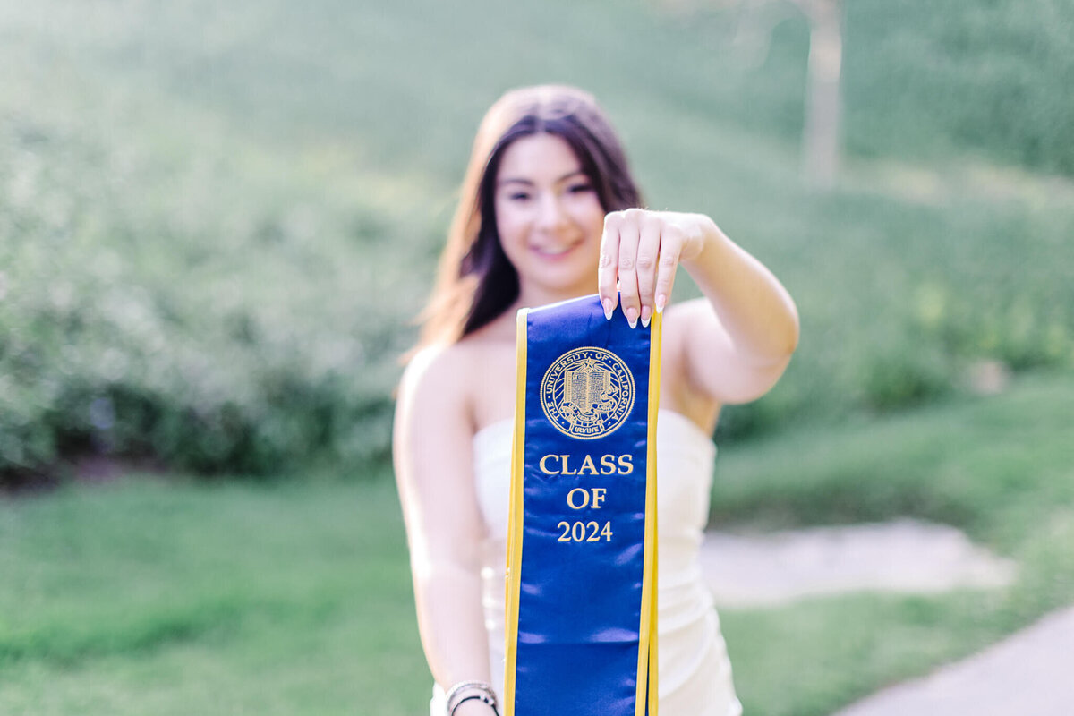 A young woman in a white dress holds a blue and yellow graduation stole with the text Class of 2024. She stands outdoors, with a grassy field in the background, smiling towards the camera.