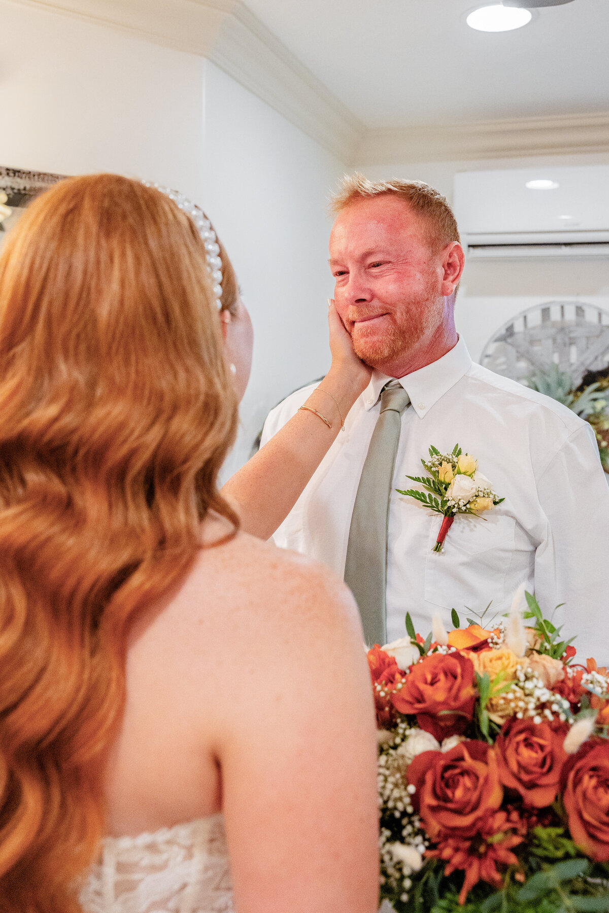 A bride gently touches her father’s face as he holds back tears, surrounded by a bouquet of autumn-colored flowers.