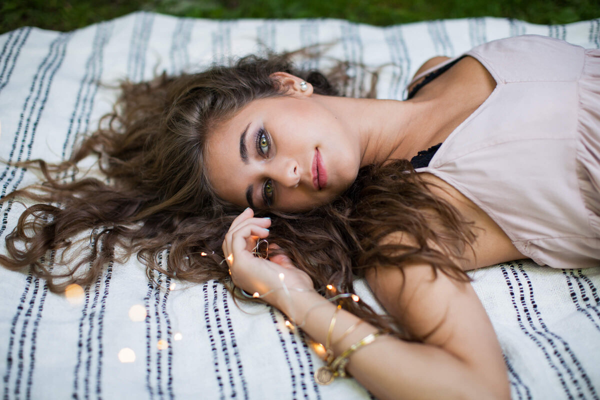 A lovely green eyed wavy haired high school senior girl lays on a blanket on the grass holding fairy lights. Photograph by Dynae Levingston