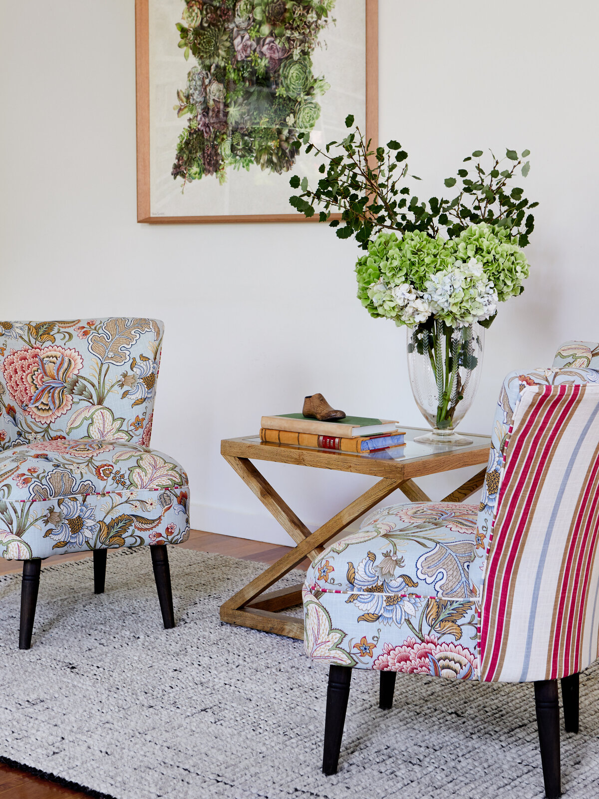 A styled sitting area in an Eynesbury home. Two armchairs with contrasting floral and striped patterns are arranged around a wooden side table, which holds a vase of hydrangeas and books.