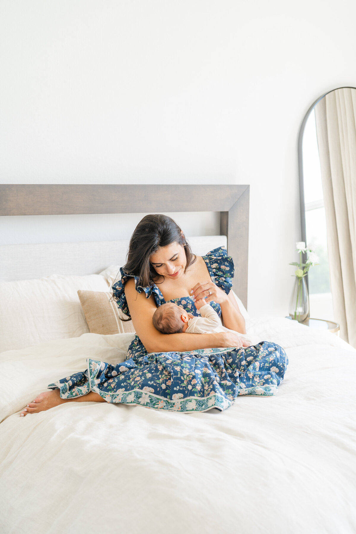 a mother sits in the center of a bed while holding her newborn. She holds his hands and looks down at him. 