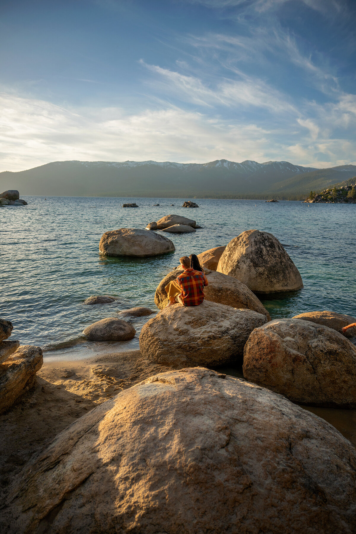 Newly engaged lovers sitting on a huge Lake Tahoe rock, looking at the snowy mountains and blue water