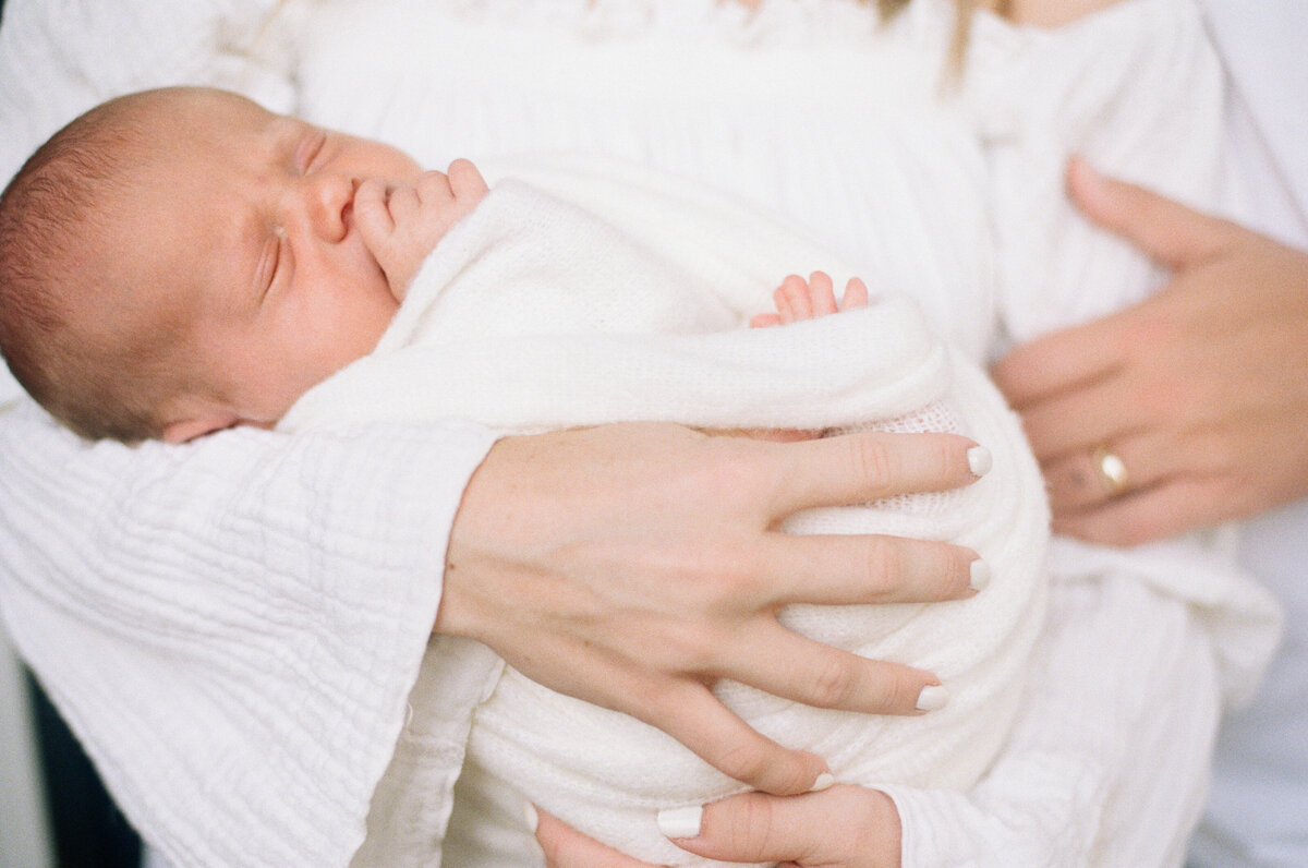Newborn portrait taken at in home in Jacksonville of baby wrapped in an ivory swaddle, sleeping in his mother’s arms