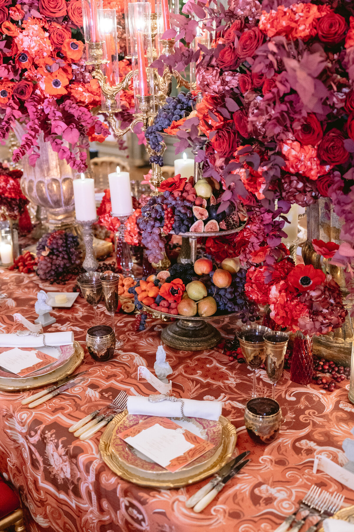 The photo was taken at Shangri La Hotel & Ritz Paris Hotel Paris, France by photographer Matteo Coltro, with Zen Film Works as Videographers. It captures a lavishly decorated table set for an event. Adorned with red and pink flower arrangements, candles, and an assortment of fruits, the table exudes an opulent and festive atmosphere. The table is covered with a patterned red tablecloth, complemented by gold-rimmed plates and elegant glassware.