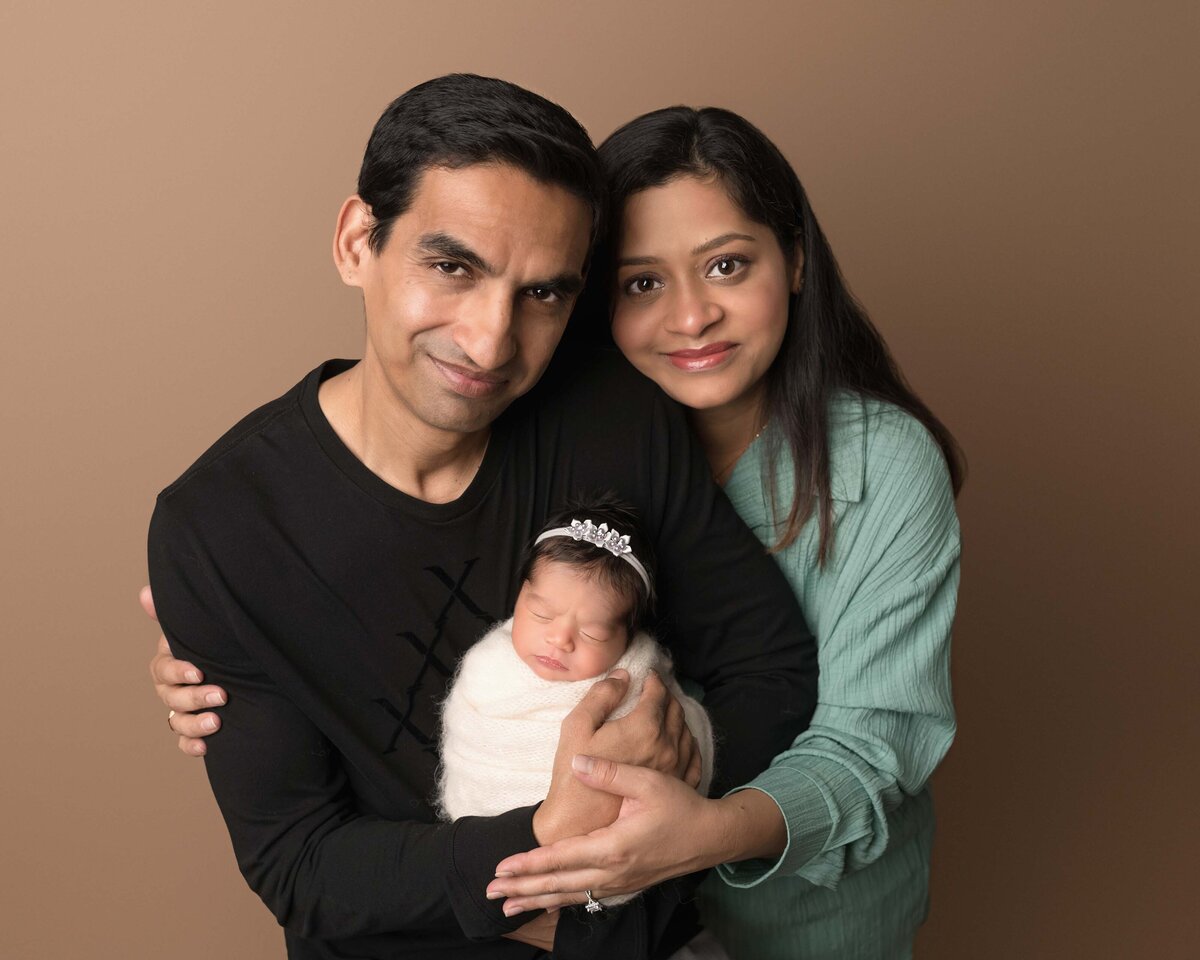 Mom and Dad pose with their newborn baby girl for their Lake Elsinore newborn photoshoot. Dad is holding baby girl and mom is standing behind dad and has her arms wrapped around dad and baby.