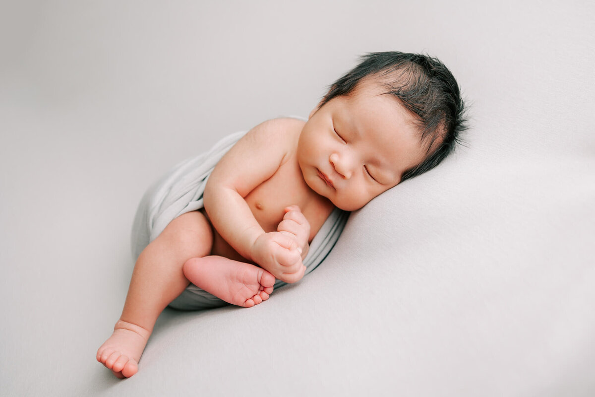 newborn boy with black hair sleeping on grey backdrop for his photography session