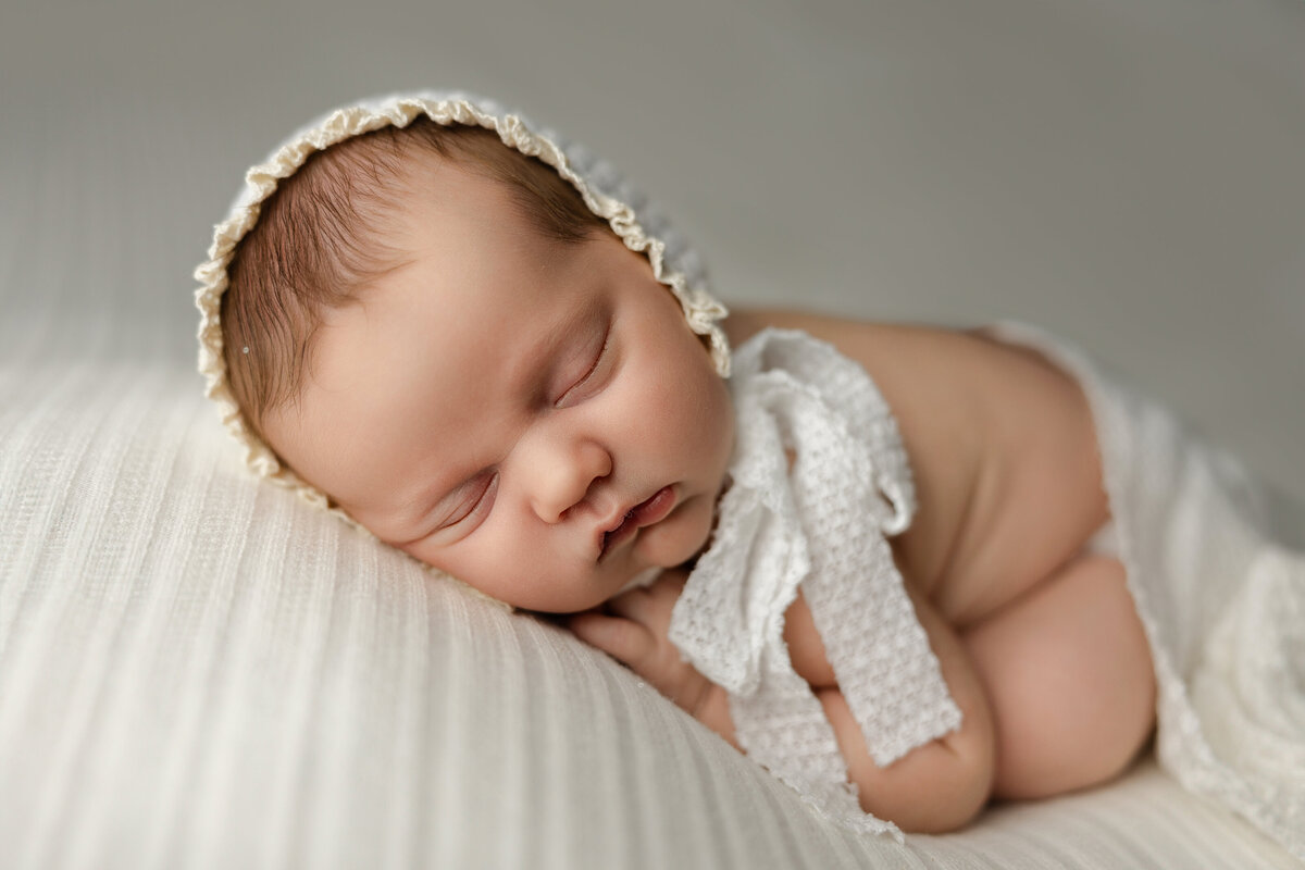 Newborn baby girl sleeping peacefully on a white textured blanket, wearing a vintage-style bonnet with delicate lace trim. Captured by Inland Empire newborn photographer Kaitlyn Dawn Photography.