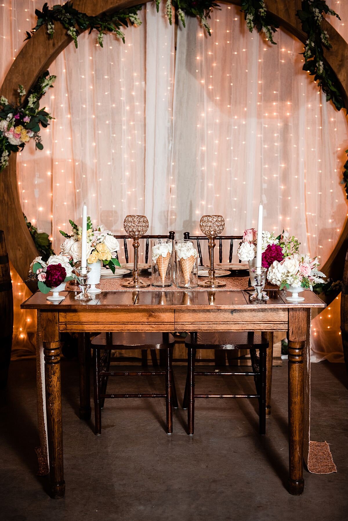 Circular dark wood ceremony arbor wrapped with a garland of greenery and ivory flowers sits against a wall with blush drapery and twinkle lights. In front of the arbor is a dark wood table decorated with rose gold crystal candle holders, long taper candlesticks, bouquets of burgundy peonies, peach roses, ivory roses, white veronica and white hydrangea. Two ice cream cones in glass vases sit at the center filled with white flowers at Grace Valley Farm.