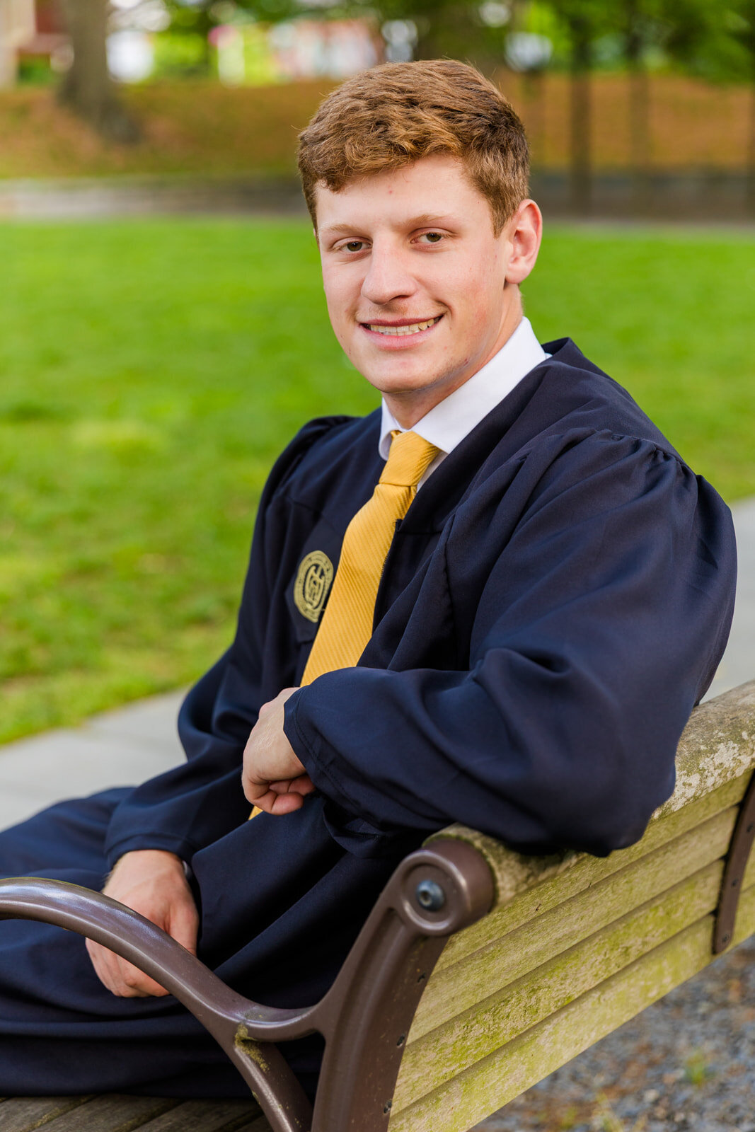 GT University senior sitting on campus on a bench looking at the camera over his shoulder during graduation portraits wearing a gown during graduation portraits with Atlanta photographer Laure Photography