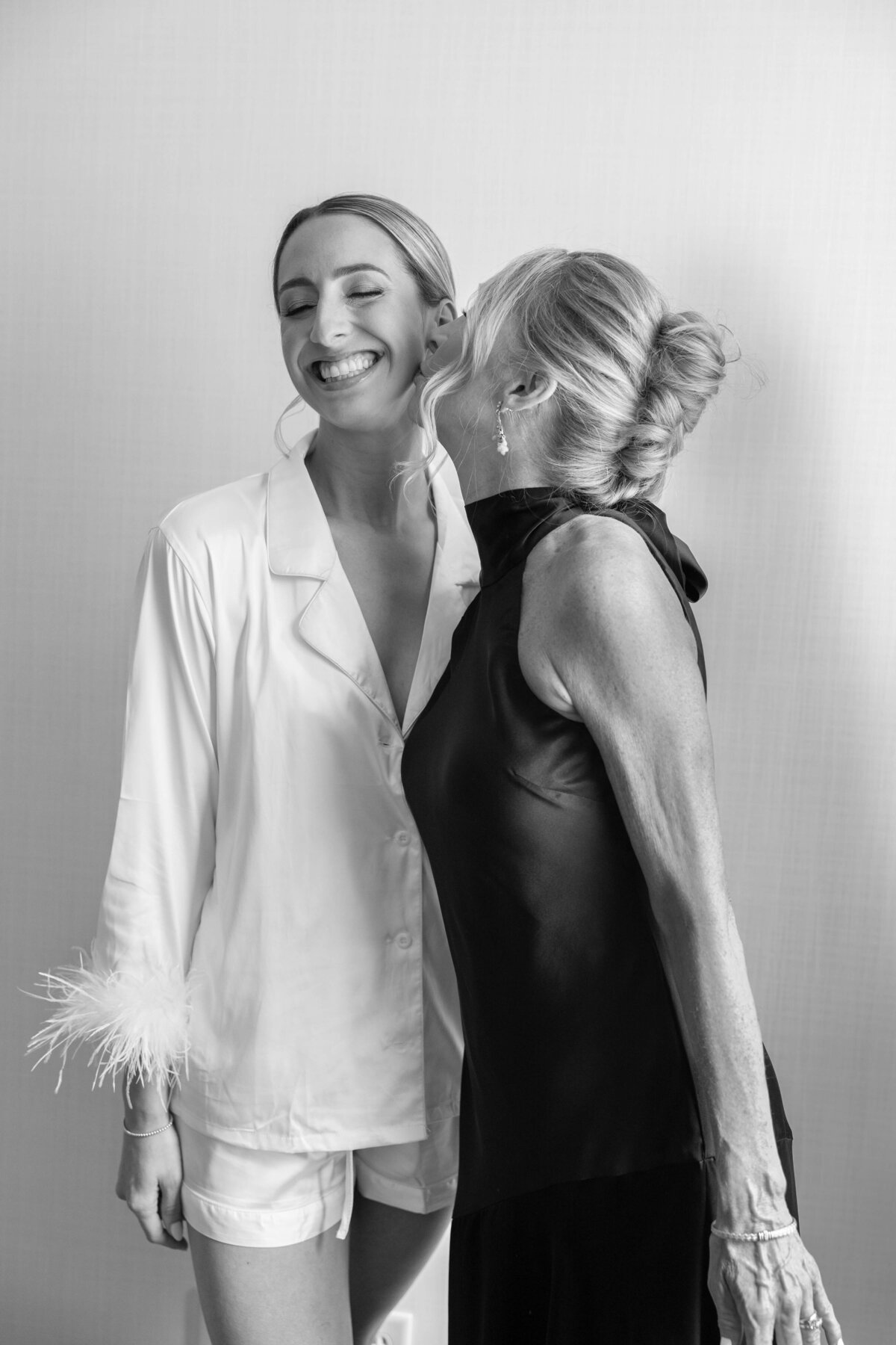 A woman in a white silk outfit smiles widely as another woman in a sleeveless black dress leans in. The scene captures a joyful, affectionate moment between them against a plain background in black and white.