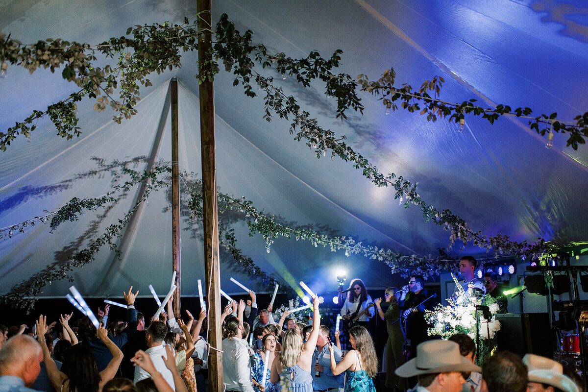  Guests celebrating on packed dance floor under greenery-draped tent ceiling with live band.