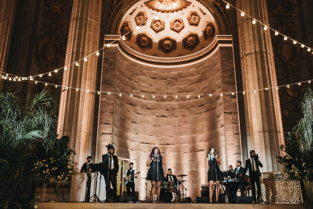This is where the photo was taken at Washington D.C., The Mellon Auditorium, one of the best luxury DC venues with European flair, captured by photographer Sarah Bradshaw. In the image, a lively band performs on an ornate stage set in an opulent hall. String lights hang above, and lush greenery decorates the sides. The backdrop is a large, elegantly carved wall with arches and intricate designs, adding to the grandeur of the setting.