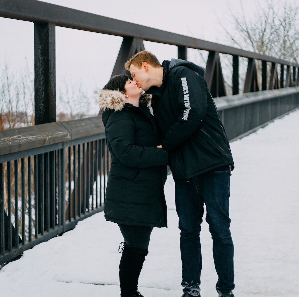 An Edmonton couple kiss on a bridge covered in snow in their black coats