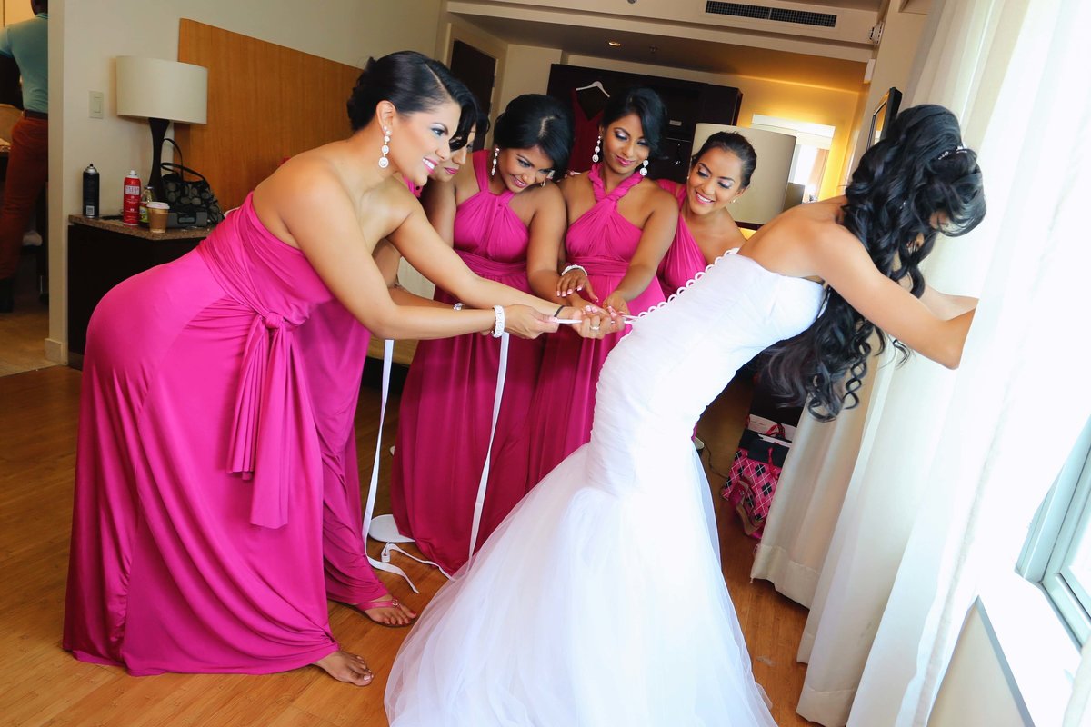 Funny shot of bride being laced into her wedding dress by bridal party wearing pink dresses. Photo by Ross Photography, Trinidad, W.I..