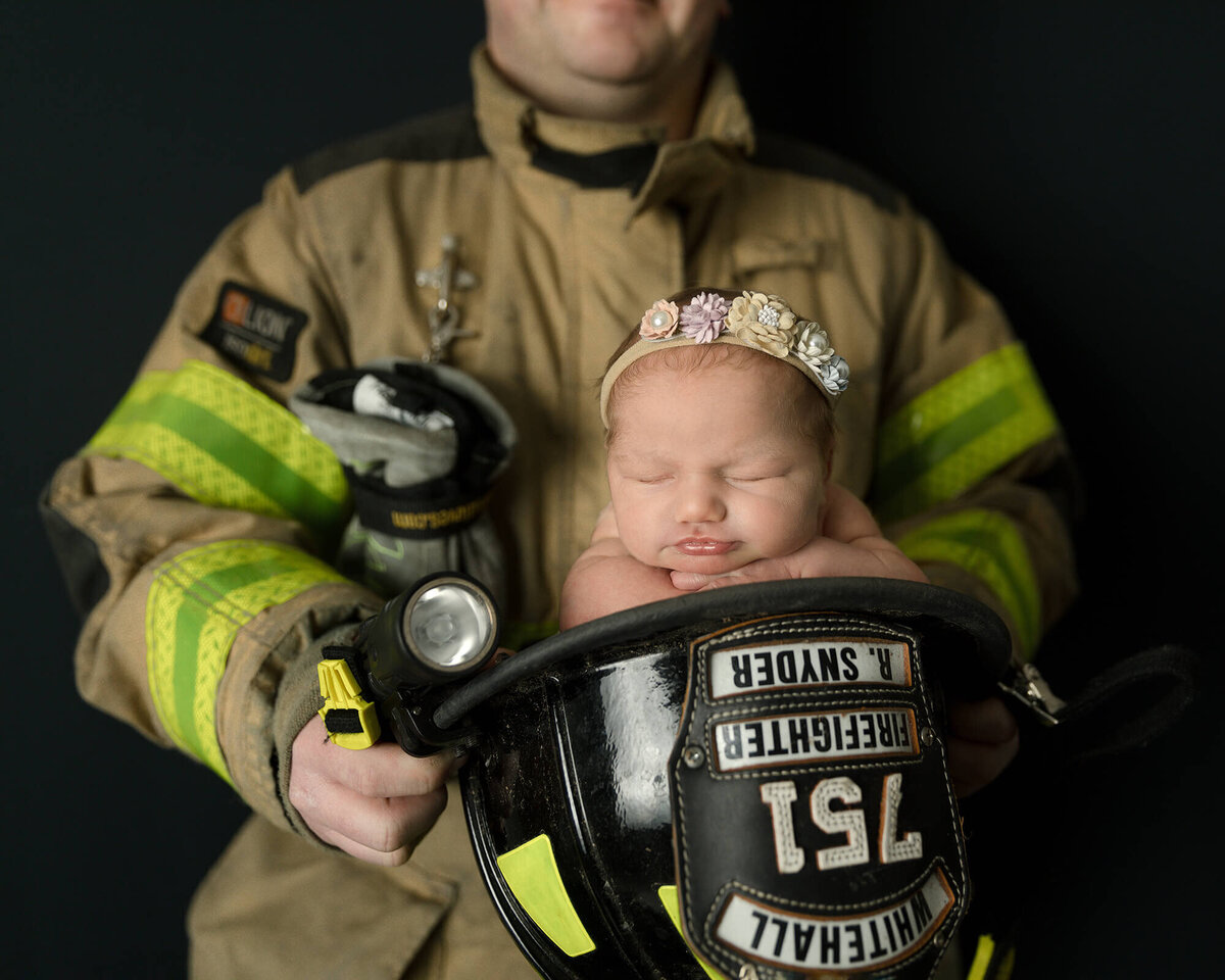 Newborn girl sleeping in her Dad's firefighter helmet while he's holding her dressed in his uniform.